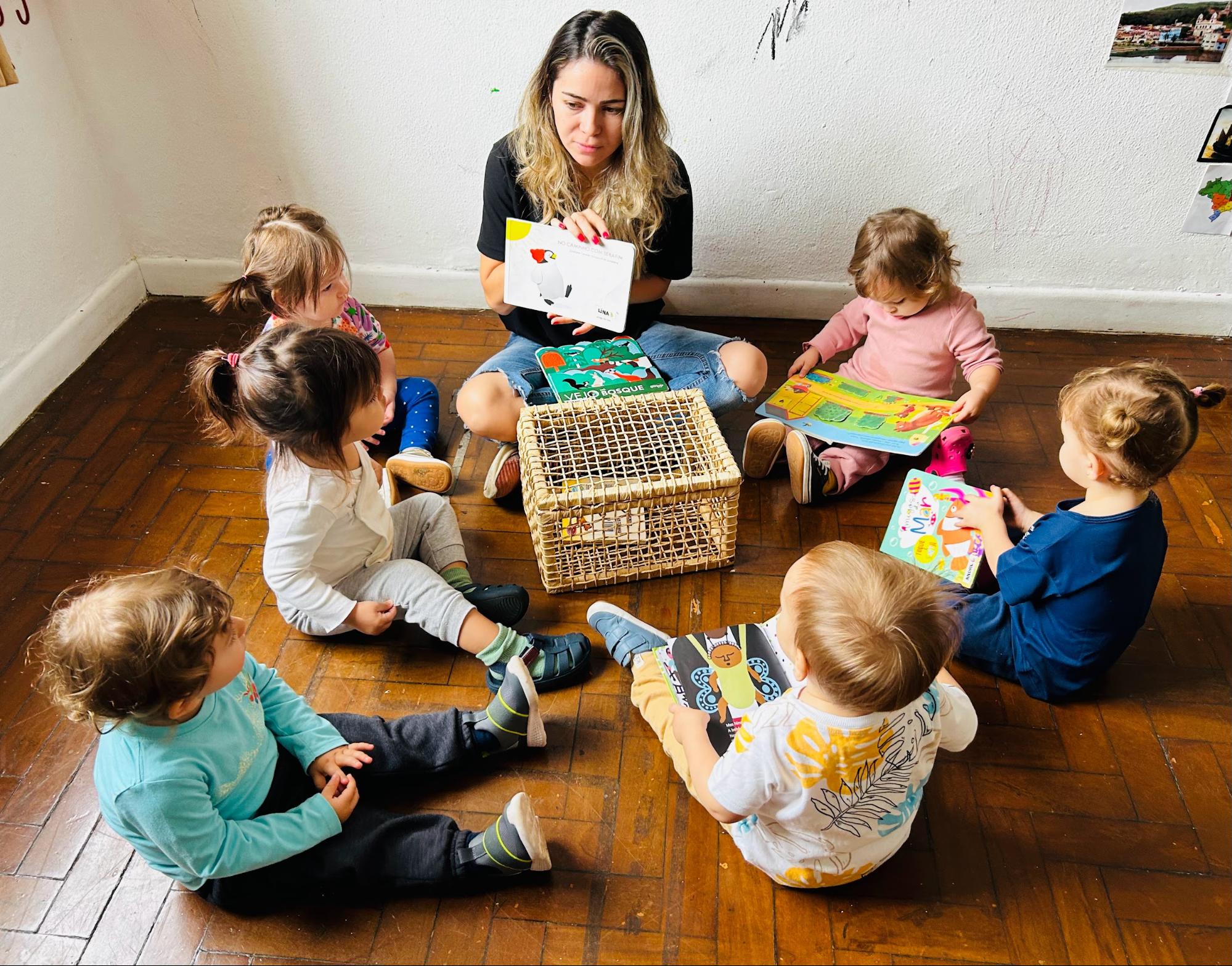 Educadora sentada em roda com bebês, todos manuseando livros ilustrados