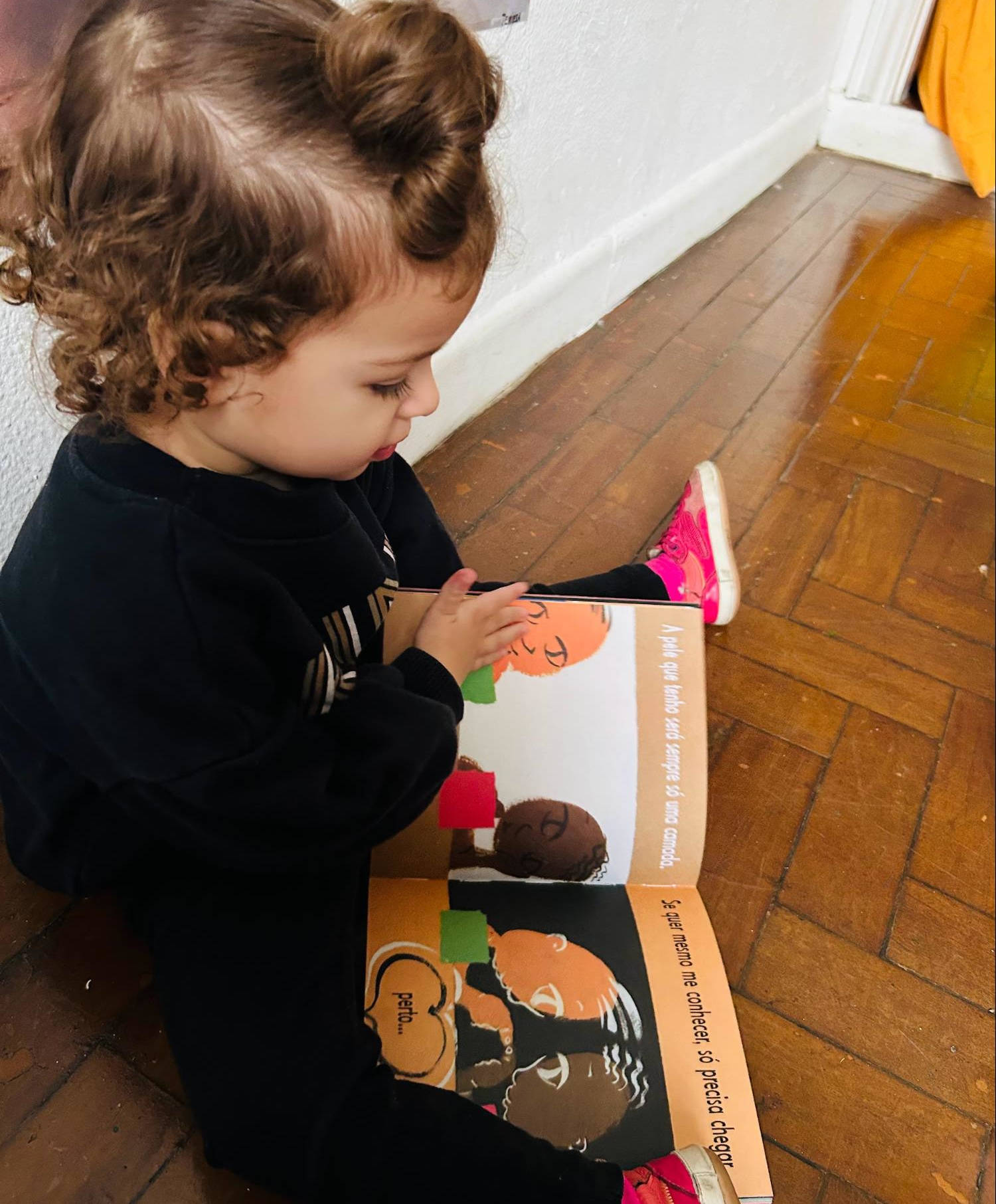 Criança sentada sozinha no chão, folheando um livro ilustrado sobre identidade e diversidade