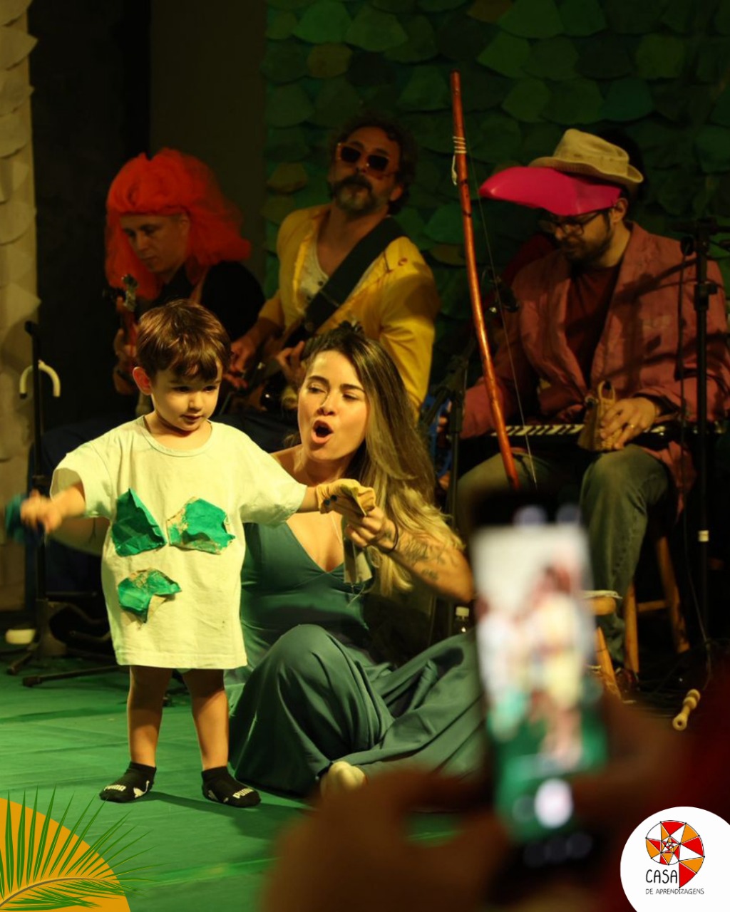 Criança pequena fantasiada cantando ao lado de uma educadora, com músicos tocando instrumentos ao fundo